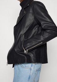 Osoba ubrana w czarną skórzaną kurtkę motocyklową z zapinanymi kieszeniami oraz jasnoniebieskie dżinsy na tle jednolitym.