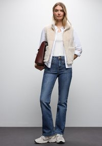 Gilet beige foderato in pelliccia sopra una camicia bianca, abbinato a jeans a zampa d'elefante blu e sneakers bianche. La modella tiene una borsa marrone. Sfondo semplice.