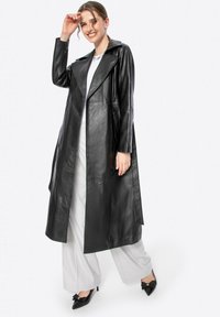 Cappotto trench in pelle nera con ampio colletto, apertura frontale, e maniche lunghe; texture liscia; design lungo fino al ginocchio. Abbinato a pantaloni chiari.