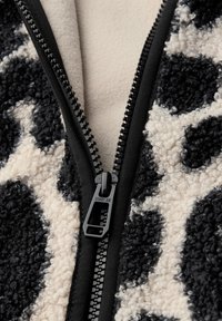 Forro polar con cremallera en negro y crema, con una superficie texturizada y esponjosa, que presenta un gran patrón de manchas y detalles en negro en la cremallera.