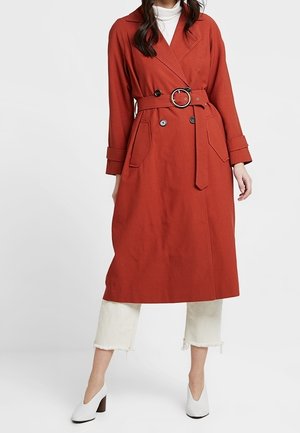 Vrouw draagt een roestkleurige trenchcoat met riem over witte cropped broek en witte schoenen met hak.