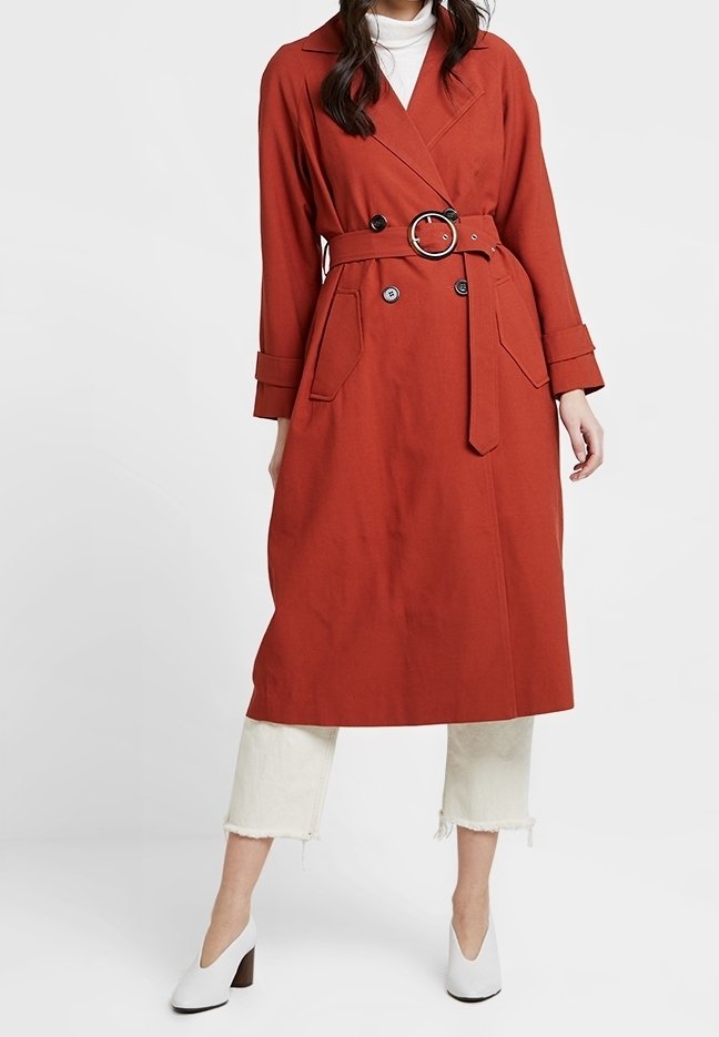 Femme portant un trench-coat rouille ceinturé par-dessus un pantalon blanc court et des chaussures blanches à talons.