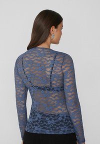 Blaues Spitzenoberteil mit langen Ärmeln, das mit floralen Mustern, durchsichtigen Materialien, einem runden Ausschnitt und einer taillierten Silhouette begeistert. Die Rückansicht hebt die filigranen Details hervor.
