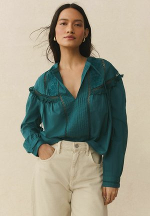 Woman wearing teal long-sleeve blouse with lace details tucked into high-waisted beige pants, standing with one hand in pocket.