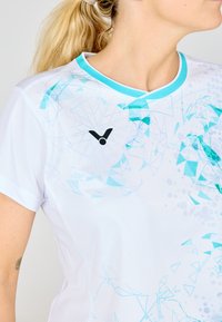 Hvid atletisk skjorte med lyseblå abstrakte geometriske mønstre; har en rund halsudskæring og et sort logo på forsiden. Glat tekstur.