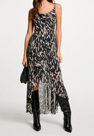 Femme portant une robe midi sans manches à motifs noirs et beiges avec un ourlet asymétrique, des bottes noires hautes jusqu’au genou et tenant un sac à main noir.