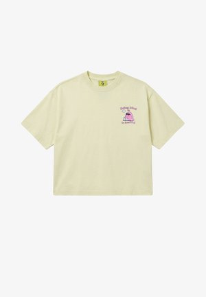Beżowy krótki crop top z krótkim rękawem z małym różowym psem z kreskówki w okularach przeciwsłonecznych oraz tekstem "Being kind is healthy" na lewej piersi.