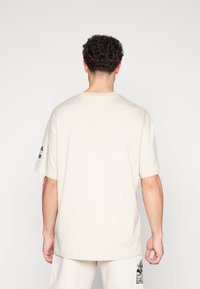 Cream-colored cotton t-shirt with short sleeves, relaxed fit, and a round neckline. Features black graphic print on the left sleeve.