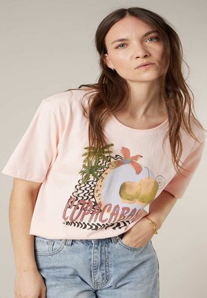 Femme aux longs cheveux bruns portant un T-shirt graphique rose clair "Copacabana" et un jean bleu clair, debout avec une main dans la poche.