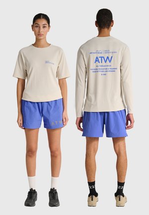 Dwa modele noszące beżowe koszule z długim i krótkim rękawem oraz niebieskie szorty, czarne i beżowe sneakersy, stoją przodem i tyłem na jednolitym tle.