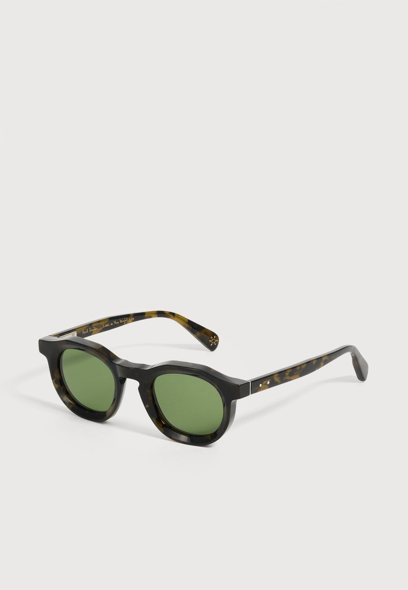 Lunettes de soleil noires à motif écaille avec des verres ronds verts et des montures épaisses, posées en biais sur une surface claire.