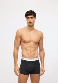 Melnas rakstūras bokseri ar baltu elastīgu jostasvietu, kurā attēlots Calvin Klein logo, pieguļošs dizains un gluda auduma tekstūra.