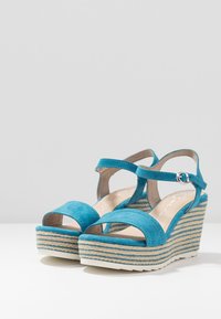 Tyrkysové semišové sandály na klínovém podpatku s překříženými popruhy a nastavitelným páskem kolem kotníku. Obsahují texturovanou tkanou platformu.