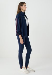 Giacca navy con zip e strisce rosse a contrasto, abbinata a leggings navy e sneaker bianche, che mette in risalto un design aderente e una texture morbida.