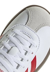 Zapatilla blanca con cuero texturizado y acentos de ante. Presenta rayas rojas, cordones blancos y una suela de goma beige. Vista en primer plano del área de los dedos.