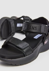 Sandalias negras con correas ajustables, hechas de tela texturizada. Presentan una suela gruesa y un panel con el logo. Diseño redondeado con plantilla acolchada.