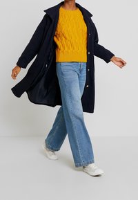 Marinblå kappa, gul kabelstickad tröja, ljusblå vida jeans och vita sneakers. Har knappar på kappan och stickad struktur på tröjan.