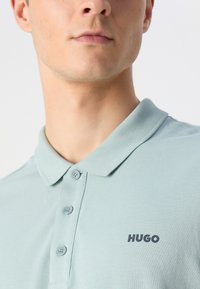 Világoszöld pólóing, texturált anyagból készült, gallérral, három gombbal és egy apró, sötétkék "HUGO" logóval a bal oldalon.