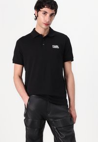 Zwart polo shirt van katoen met een klassieke kraag, korte mouwen en een wit logo aan de linkerkant. Gecombineerd met zwarte leren broek.