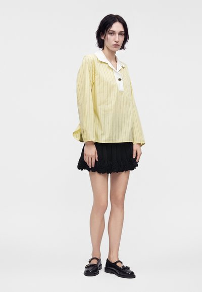 Femme aux cheveux courts et foncés portant un chemisier jaune rayé à manches longues, une mini-jupe noire à volants et des chaussures Mary Jane noires, debout devant un fond uni.