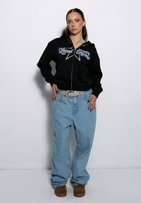 Czarna bluza z kapturem na zamek, zdobiona kryształkami i białym napisem, zestawiona z jasnoniebieskimi luźnymi dżinsami oraz brązowym obuwiem.