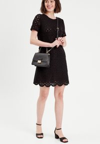 Robe en dentelle noire à manches courtes, ourlet festonné, motifs floraux ; associée à un sac à main noir et des sandales à talons.