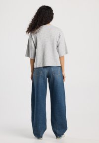 Woman with curly hair wearing a loose gray short-sleeve top and wide-leg blue jeans, standing facing away on a plain background.