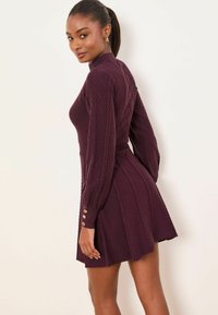 Gebreide jurk in diep bordeauxrood met lange mouwen, ruitpatronen en knoopaccenten bij de manchetten. Getailleerd lijfje dat uitloopt in een rok.