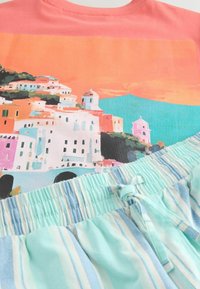 Chemise corail imprimée avec un motif de ville côtière pittoresque, associée à un short à taille élastique rayé bleu clair et blanc, orné d'un petit nœud à l'avant.