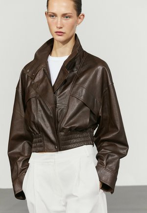 Junge Frau trägt eine dunkelbraune, kurzes Lederjacke mit hohem Kragen und weiße, hoch taillierte Hose, steht vor einfarbigem Hintergrund.