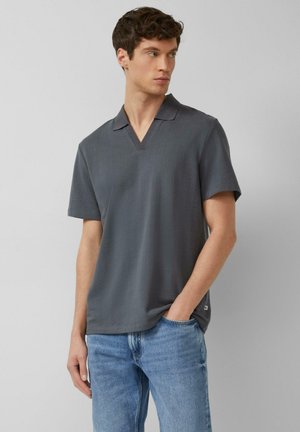 Młody mężczyzna w ciemnoszarej koszulce polo z krótkim rękawem i niebieskich dżinsach, stoi z jedną ręką w kieszeni, patrząc na bok.