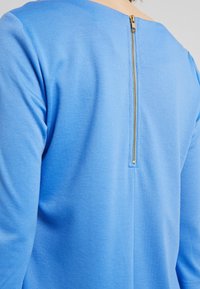 Blauwe langemouw top met ronde halslijn en een verborgen gouden rits aan de achterkant, gemaakt van zachte, gestructureerde stof.