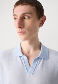 Világoskék pólóing texturált anyagból, rövid gallérral és részleges gombolással, semleges háttér előtt elhelyezve.