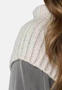 Spalla di un maglione lavorato a maglia in off-white con coste texturizzate. Tessuto grigio chiaro visibile sotto. Aspetto liscio e accogliente.