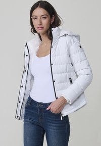 Chaqueta acolchada blanca con cierre de cremallera y botones, capucha y detalles en negro. Llevada sobre una camisa blanca y combinada con jeans azules.