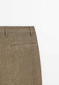Gros plan sur la partie supérieure d'un pantalon marron texturé présentant des passants de ceinture et une unique poche passepoilée à l'arrière.
