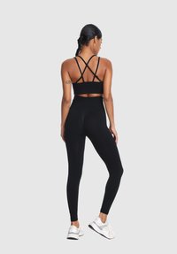 Žena s preplietanými vlasmi oblečená v čiernom športovom podprsenke s krížovým dizajnom, vysokých legínach a bielych teniskách, stojí na pozadí bez vzoru.