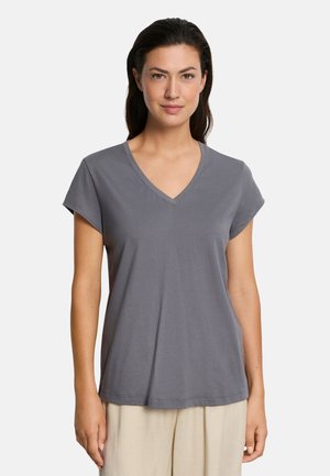 Kobieta w szarej koszulce z krótkim rękawem i dekoltem w serek oraz jasnych spodniach, stojąca na tle jednolitego białego tła.