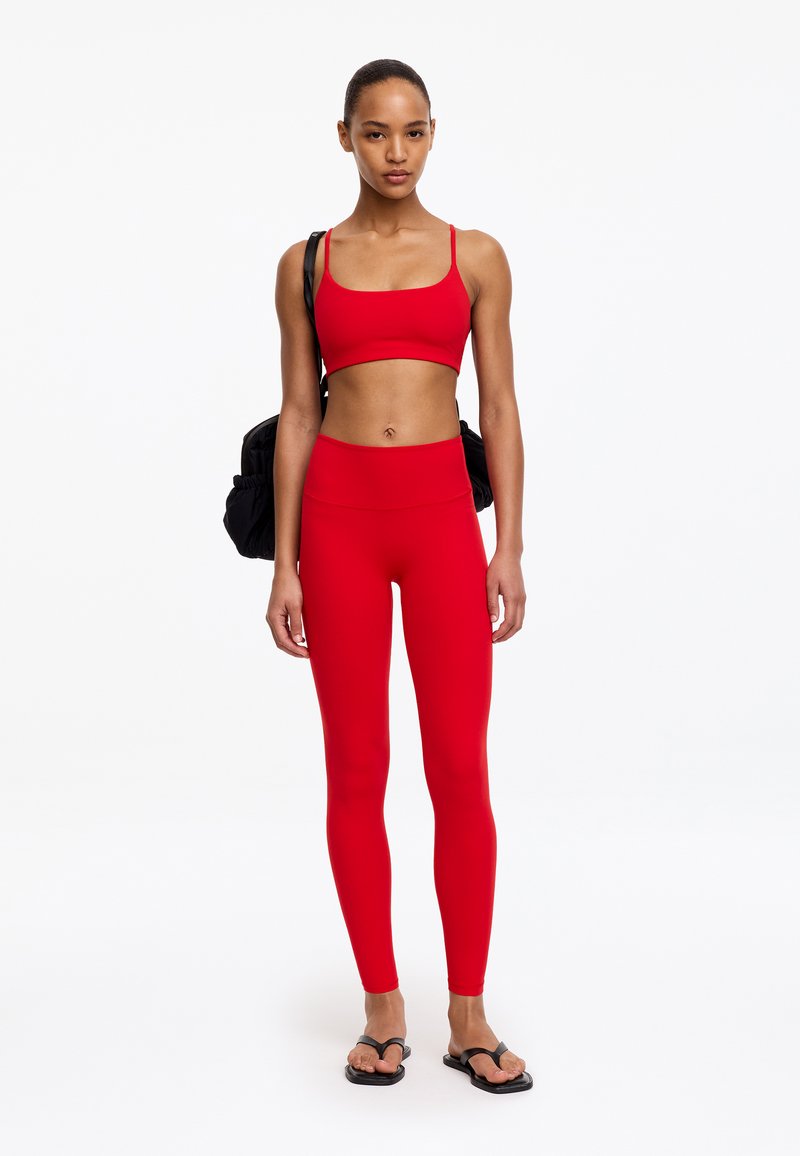 Jeune femme debout portant un brassière de sport rouge et des leggings avec un sac à dos noir et des sandales noires sur fond blanc.
