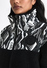 Black and gray patterned jacket with a high collar and textured fleece lower section. Features a round logo patch on the chest.
