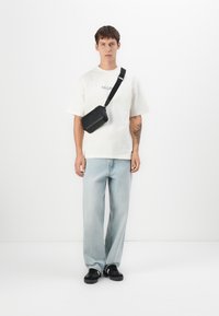 Young man wearing white graphic t-shirt, light blue jeans, black sneakers, and a black crossbody bag against a plain white background.