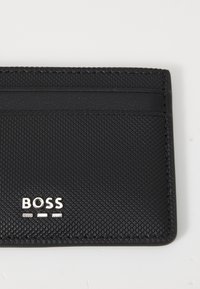 Melnā kartes turētājs, izgatavots no tekstūrēta sintētiskā materiāla. Tam ir vairāki kartes nodalījumi un sudraba "BOSS" logo priekšpusē.