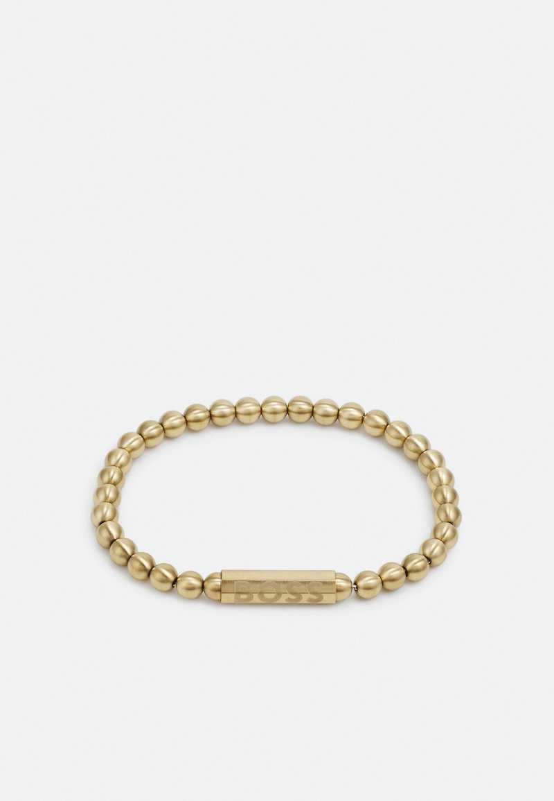 Pulsera elástica en tono dorado que presenta cuentas esféricas lisas y un acento rectangular plano y grabado con el nombre de la marca.