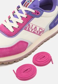 Sneaker viola e rosa con una tomaia in suede e tessuto, dettagli di branding, accenti crema e due lacci rosa di ricambio accanto ad essa.