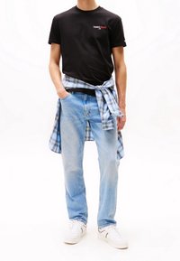 Czarna bawełniana koszulka z logo "TOMMY JEANS", jasnoniebieskie dżinsy oraz koszula w kratę związana w talii. Białe sneakersy dopełniają stylizację.