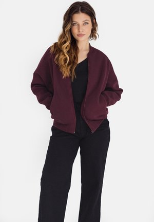 Kobieta z długimi falującymi włosami, ubrana w ciemnoczerwoną bomberkę na czarną bluzkę i czarne spodnie, stoi z rękami w kieszeniach kurtki.