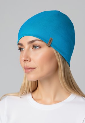 Niebieska dzianinowa czapka z gładką teksturą i dopasowanym krojem. Posiada małą brązową metkę z boku. Noszona z białym topem.