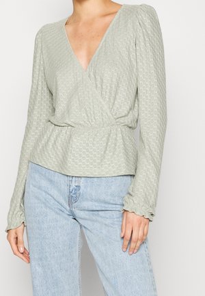 Vrouw draagt een lichtgroene, getextureerde blouse met lange mouwen, een wikkelmodel met een elastische taille en gerimpelde manchetten, gecombineerd met lichtblauwe jeans met hoge taille.