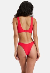 Roter Bikini bestehend aus einem strukturierten Stoff, einem Top mit weitem Rückenausschnitt und breiten Trägern sowie hochgeschnittenen Bikinihosen. Minimale Details und klare Linien.