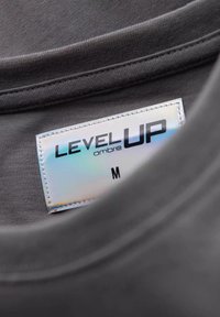 Šedá tkanina trička s hladkou strukturou, s holografickou etiketou, na které je napsáno "LEVEL UP ombre M." Viditelné švy na límci.